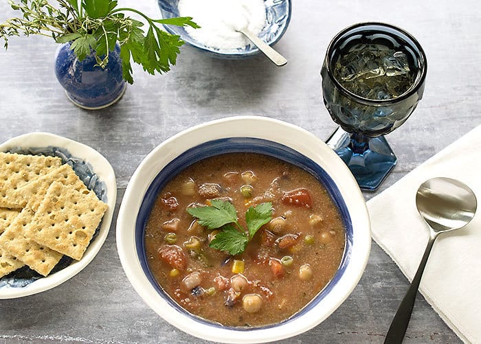 Instant Pot Teff Vegetable Soup in a ceramic bowl, iced tea in a blue glass, and crackers on a side plate