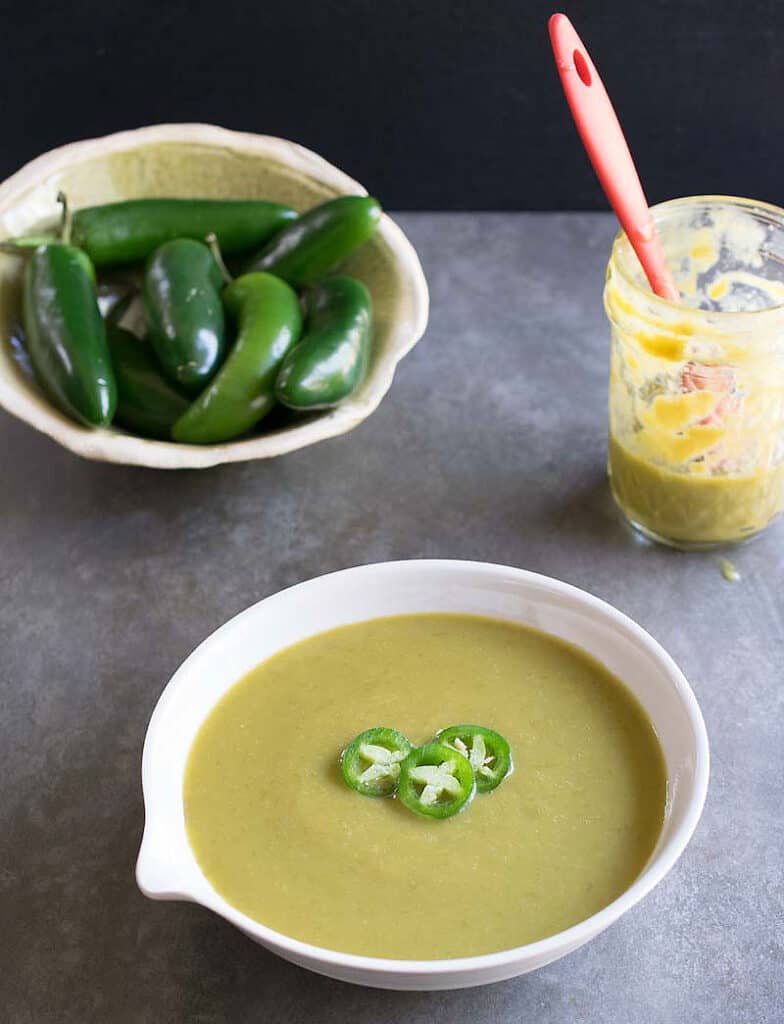 Photo with a jar of Jalapeno Hot Sauce, a handmade bowl of fresh jalepenos, and a white bowl with a pour spout full of Jalapeno Hot Sauce.