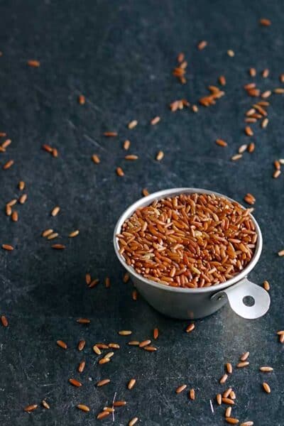 Uncooked whole grain red rice in a tin measuring cup with grains scattered on a table.