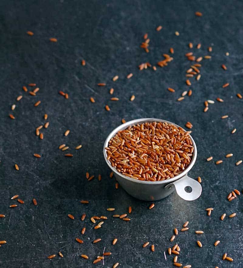 Uncooked whole grain red rice in a tin measuring cup with grains scattered on a table.