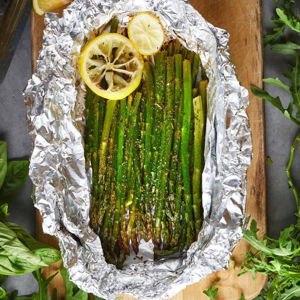 Asparagus spears with lemon slices in a foil packets on a wooden cutting board.